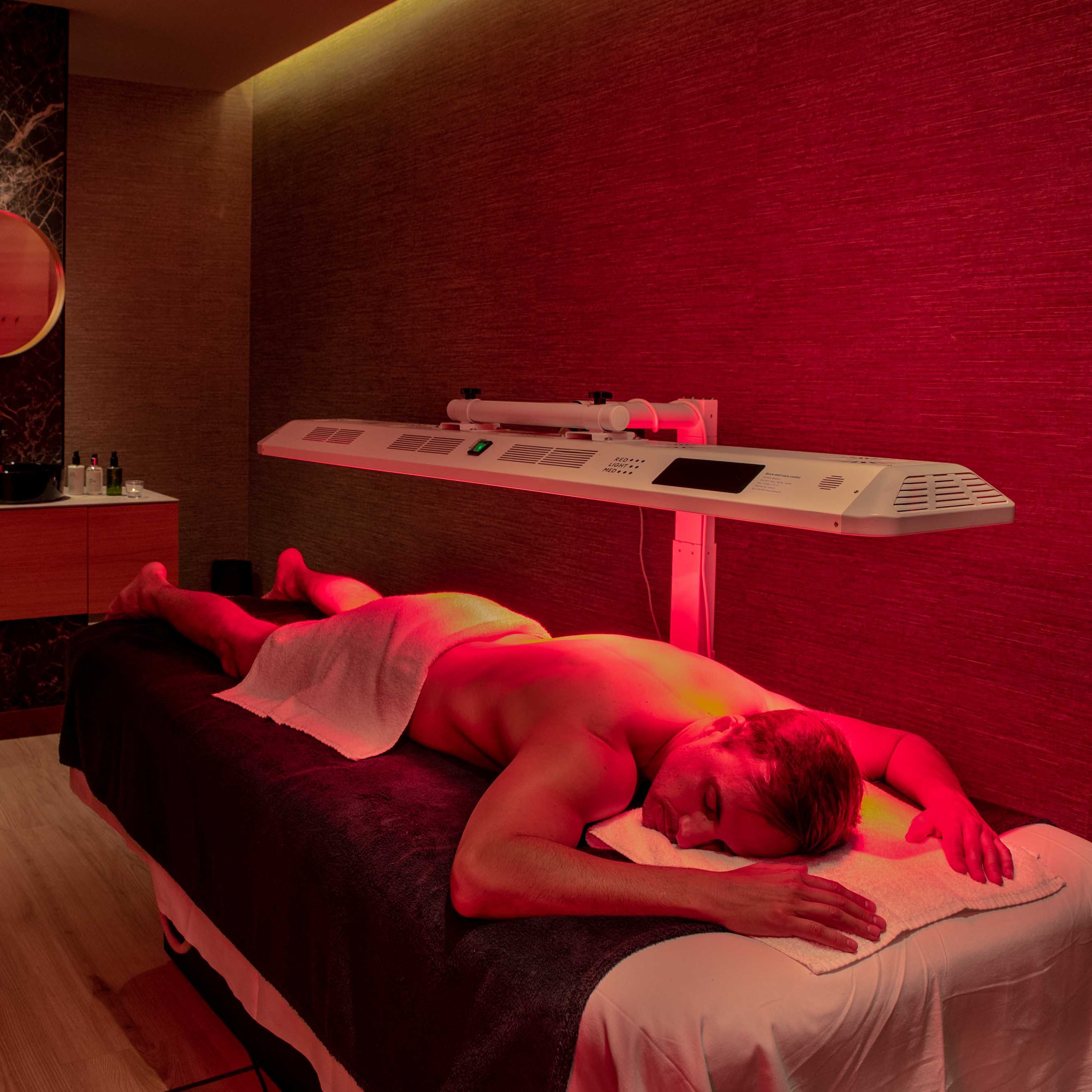 A man lies face down on a massage table in a spa room under warm red ambient lighting, with a white towel wrapped around his lower body. A long white treatment table and control panel are visible against a textured red wall, with wooden cabinets and a marble backsplash in the background.